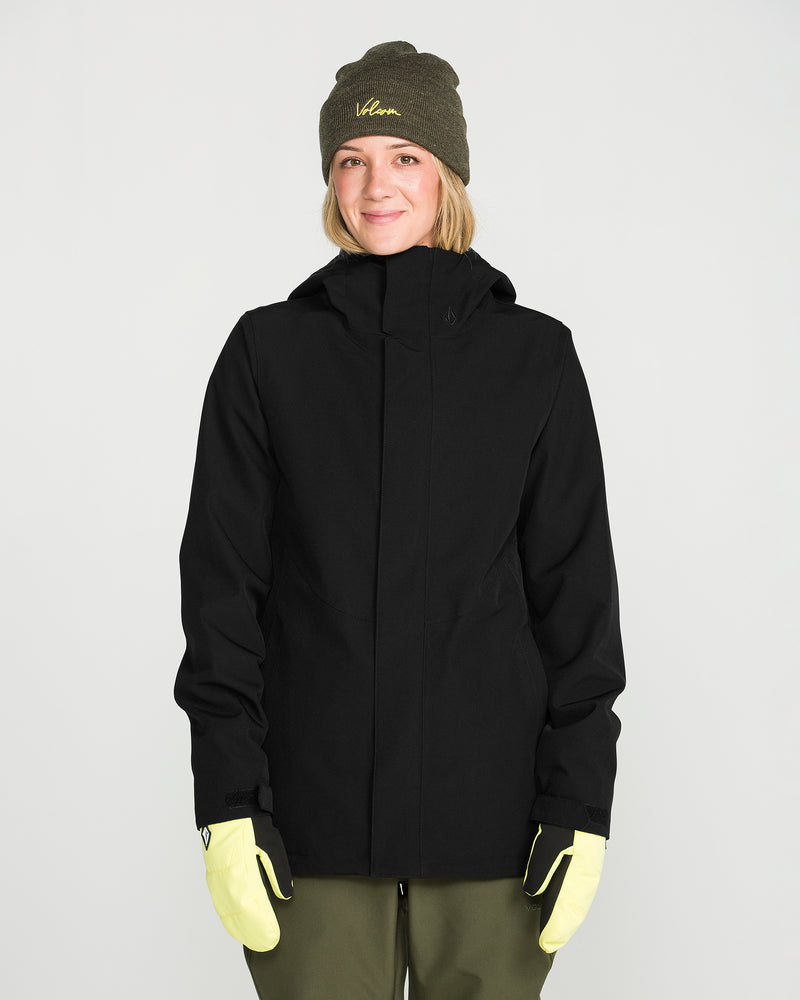 A person wearing the Volcom Women’s Stoney Shadow Insulated Jacket in black, paired with an olive green beanie, olive pants, and light yellow gloves, stands smiling against a plain white background.