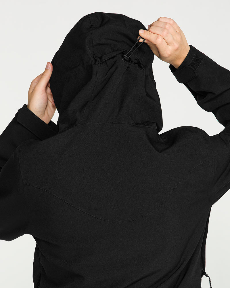 A person adjusts the drawstring on the hood of a black Volcom Women’s Stoney Shadow Insulated Jacket. The image, taken from behind, showcases their hands and upper back against a plain background, emphasizing the sleek design and 15K waterproof rating of this women’s snowboard jacket.