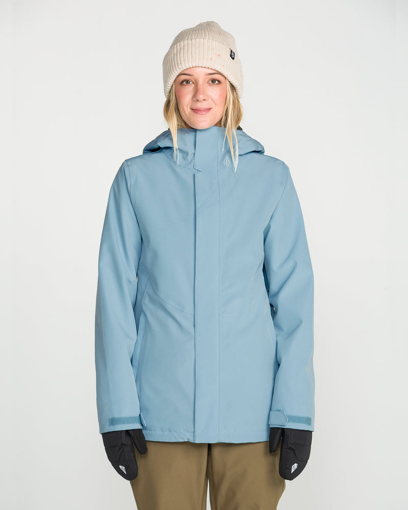 A person wearing the Volcom Women’s Stoney Shadow Insulated Jacket in light blue, styled with a beige knit beanie, black gloves, and olive green pants, stands facing the camera against a plain white background.