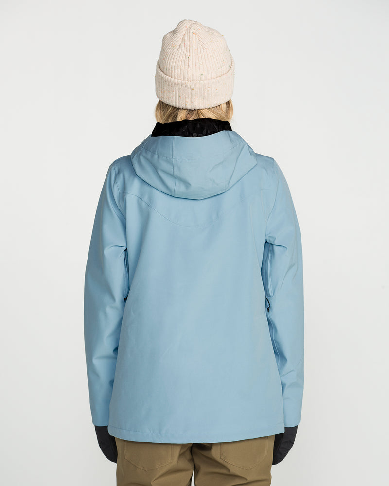 A person facing away, wearing the Volcom Women’s Stoney Shadow Insulated Jacket in light blue with recycled insulation, paired with a beige knit beanie, black gloves, and tan pants against a plain white background.