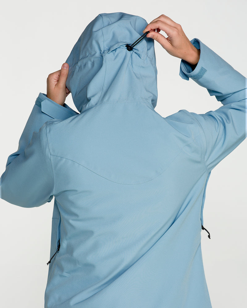 A woman is seen from the back adjusting the hood drawstring of the Volcom Women’s Stoney Shadow Insulated Jacket in light blue. The waterproof snowboard jacket features recycled insulation, and is set against a plain, neutral background.