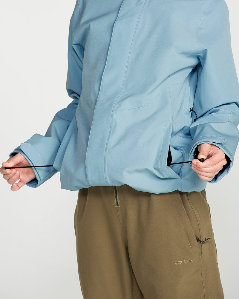 A person wearing the Volcom Women’s Stoney Shadow Insulated Jacket in light blue with recycled insulation and olive green pants adjusts the fit by pulling the jacket’s hem drawstring.