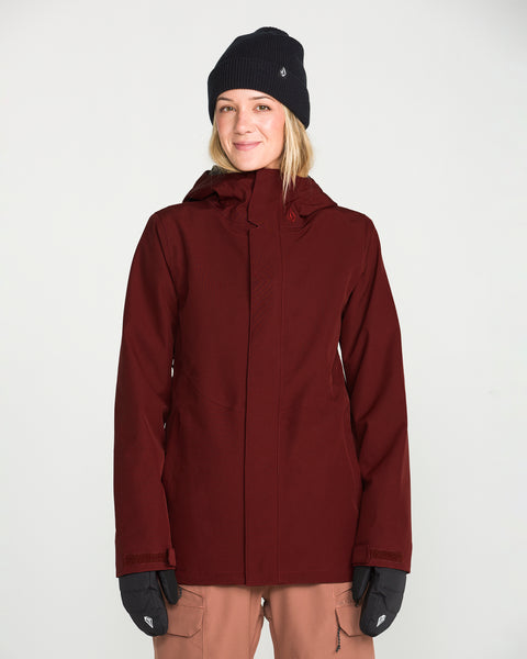 A woman wearing the Volcom Women’s Stoney Shadow Insulated Jacket in maroon, paired with a black beanie and black gloves, smiles at the camera against a plain white background.