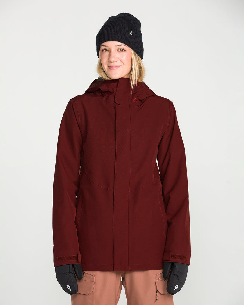 A woman wearing the Volcom Women’s Stoney Shadow Insulated Jacket in maroon, paired with a black beanie and black gloves, smiles at the camera against a plain white background.
