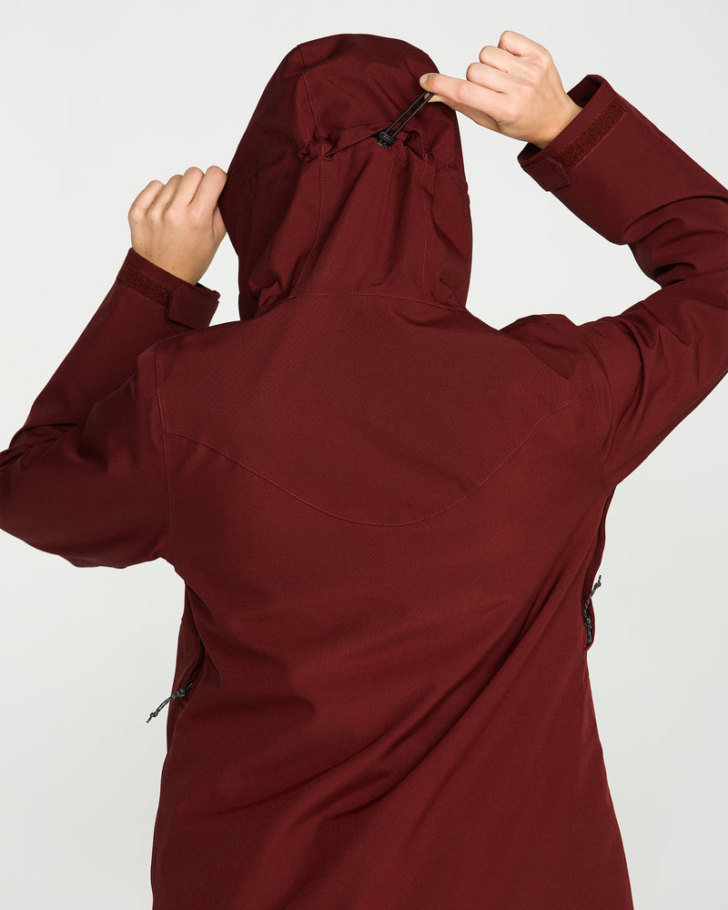 A person wearing the Volcom Women’s Stoney Shadow Insulated Jacket in maroon stands facing away, adjusting the hood with one hand while holding its edge with the other; the background is plain and light-colored.