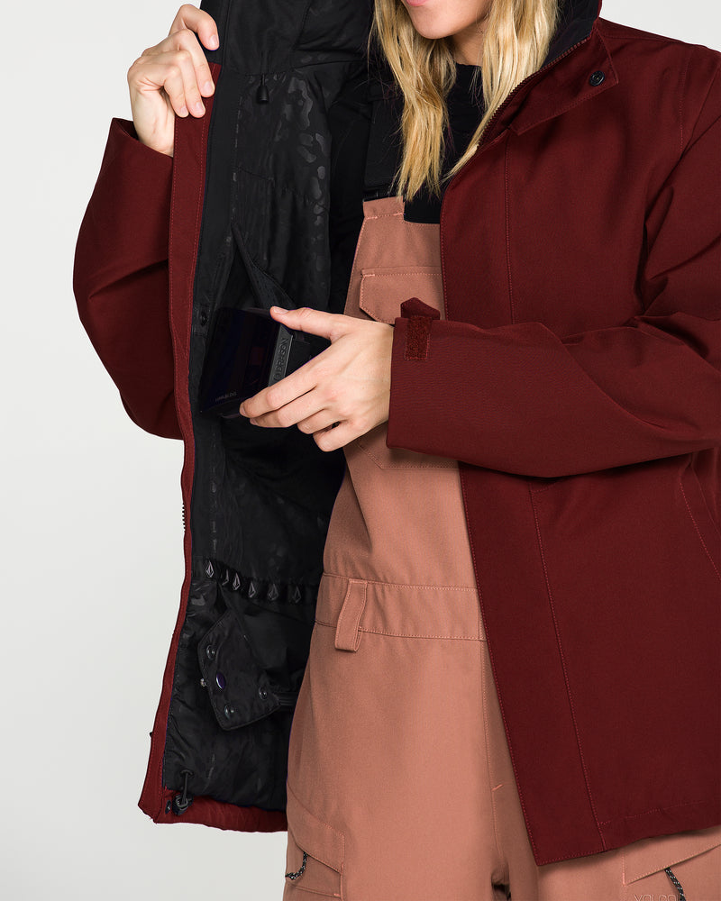 Wearing the Volcom Women’s Stoney Shadow Insulated Jacket in maroon, a person with their face partially out of frame slips a phone into the inner pocket of their open jacket while also wearing tan overalls.