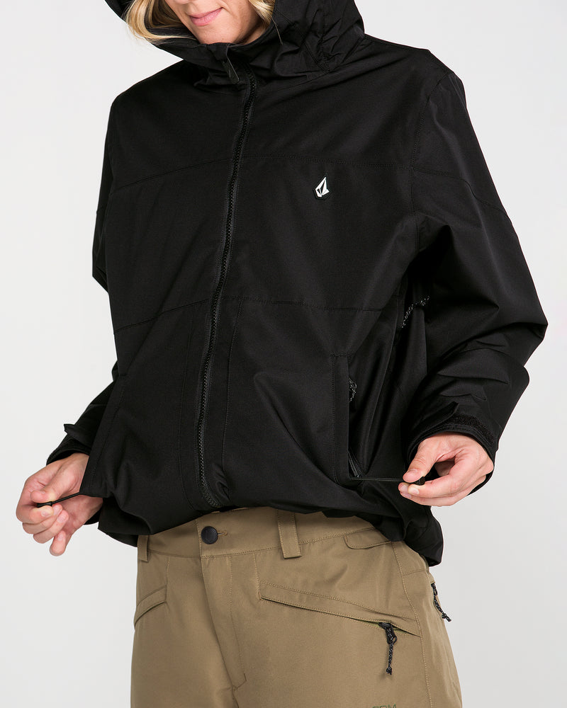 A person, with only the lower face visible, is wearing the Volcom Women’s Steele Insulated Jacket—an insulated black ski jacket featuring a small white Volcom logo on the chest, a hood, and eco-friendly recycled insulation for warmth—paired with tan pants and holding the bottom edge of the jacket.