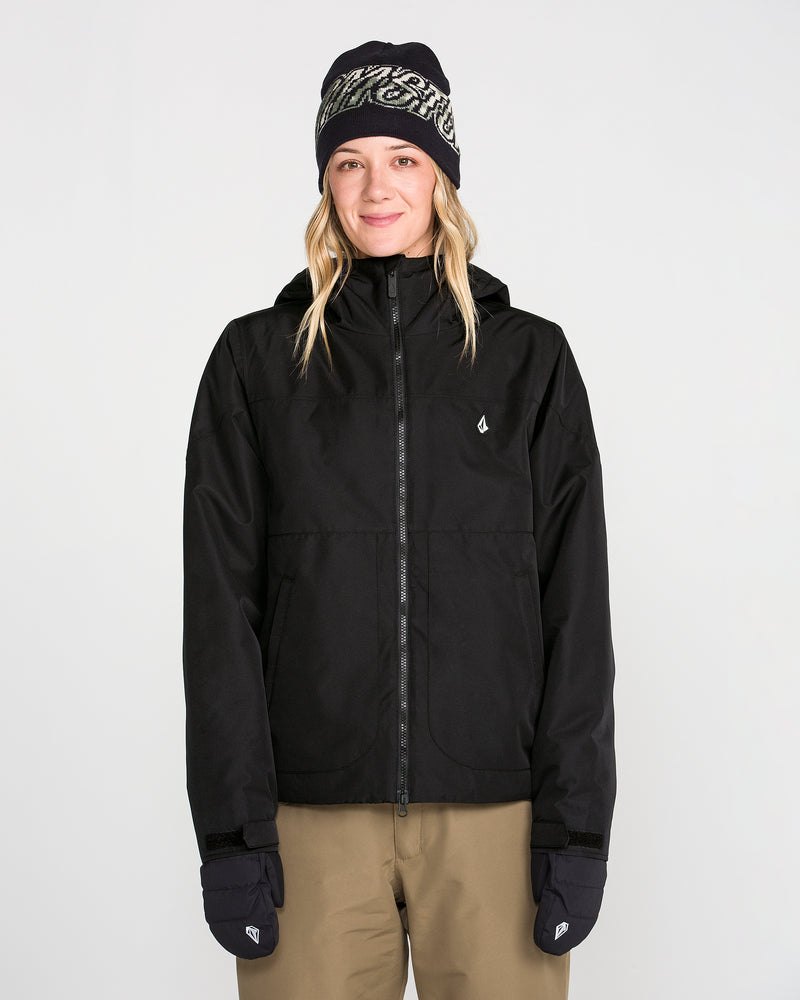 A person stands facing forward against a plain white background, wearing the Volcom Women’s Steele Insulated Jacket in black with recycled insulation, paired with a patterned black beanie, black gloves, and tan pants.