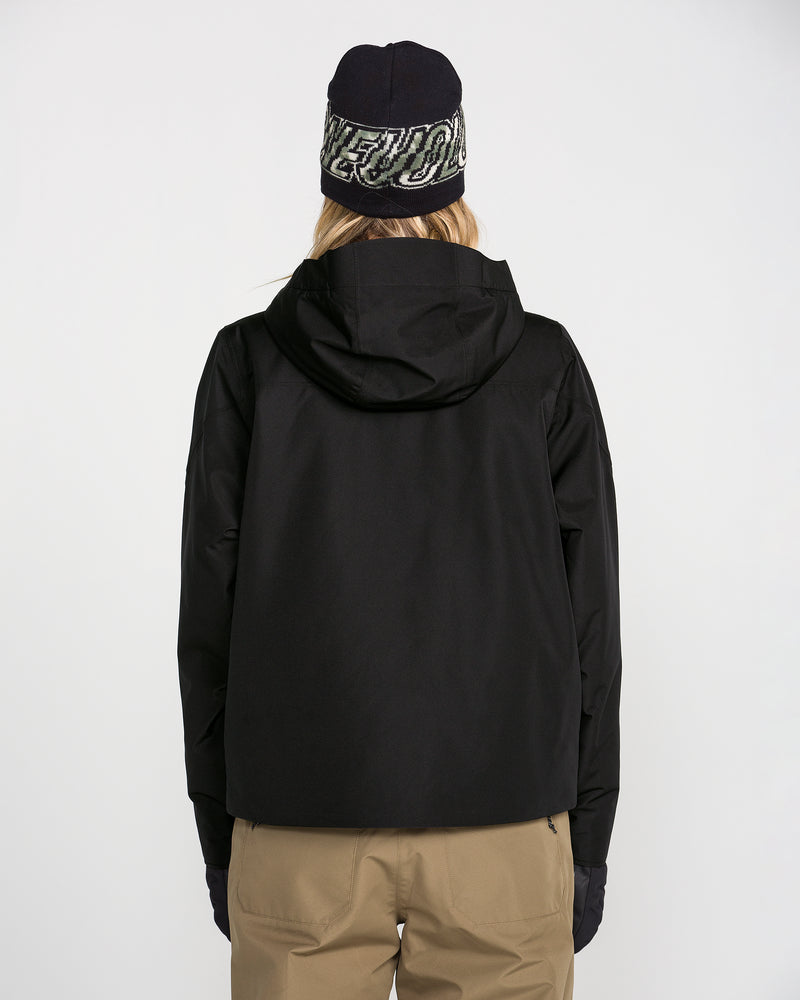 A person is shown from the back against a plain white background wearing the Volcom Women’s Steele Insulated Jacket in black, which features recycled insulation, along with a navy beanie with white text, tan pants, and black gloves.