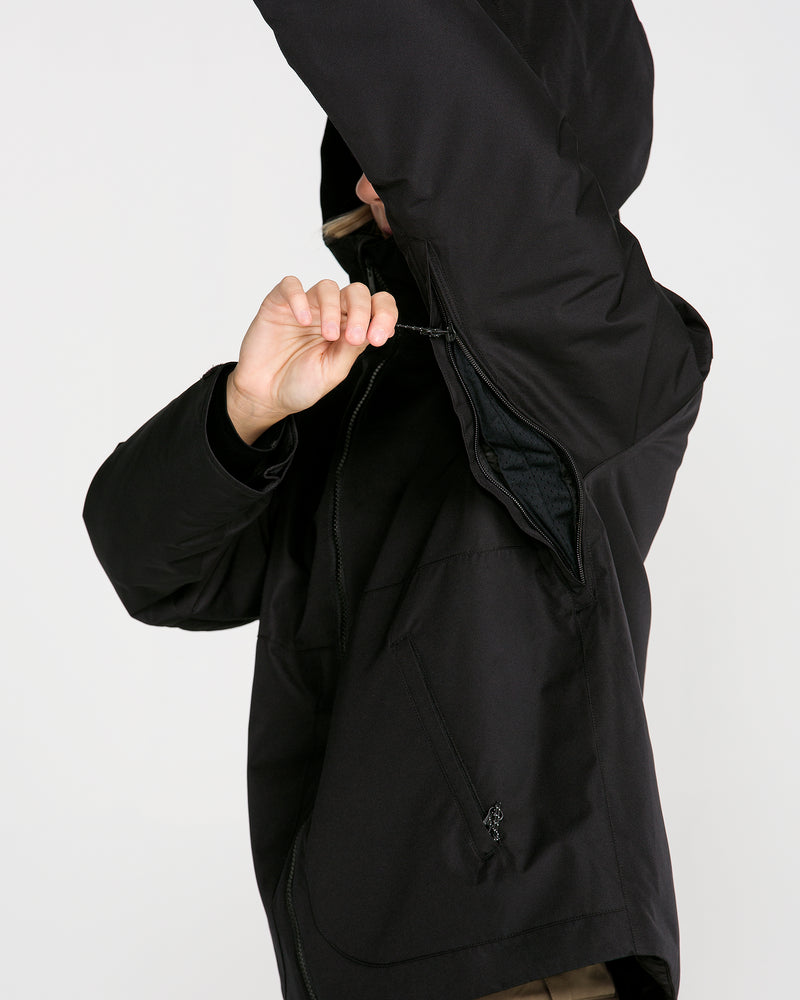 A woman in the Volcom Women’s Steele Insulated Jacket unzips an underarm vent, highlighting the jacket’s breathable design and recycled insulation, set against a plain light background.