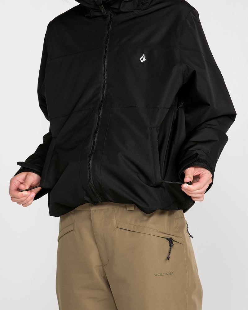 A person is wearing the Volcom Women’s Steele Insulated Jacket in black, featuring a small white logo and recycled insulation for added warmth, paired with tan pants that have a zipper pocket. The face is not visible; only the torso and hips are shown.