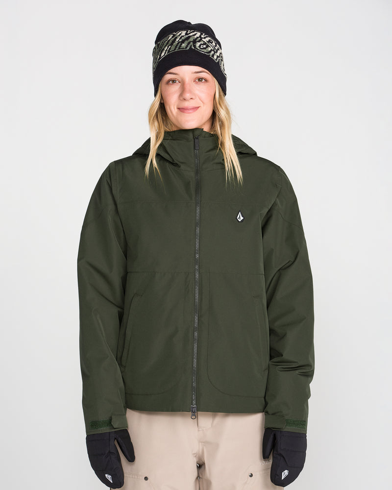 A person is wearing the Volcom Women’s Steele Insulated Jacket in dark green, paired with beige pants, black gloves, and a patterned black beanie, standing facing forward against a plain light background.