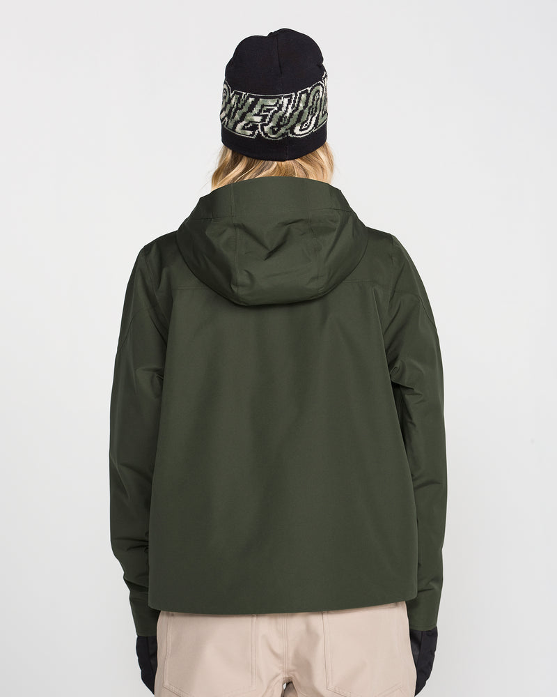 A person is facing away from the camera against a plain white background, wearing the Volcom Women’s Steele Insulated Jacket in dark green, paired with black gloves, beige pants, and a black beanie with white text. The jacket features recycled insulation.