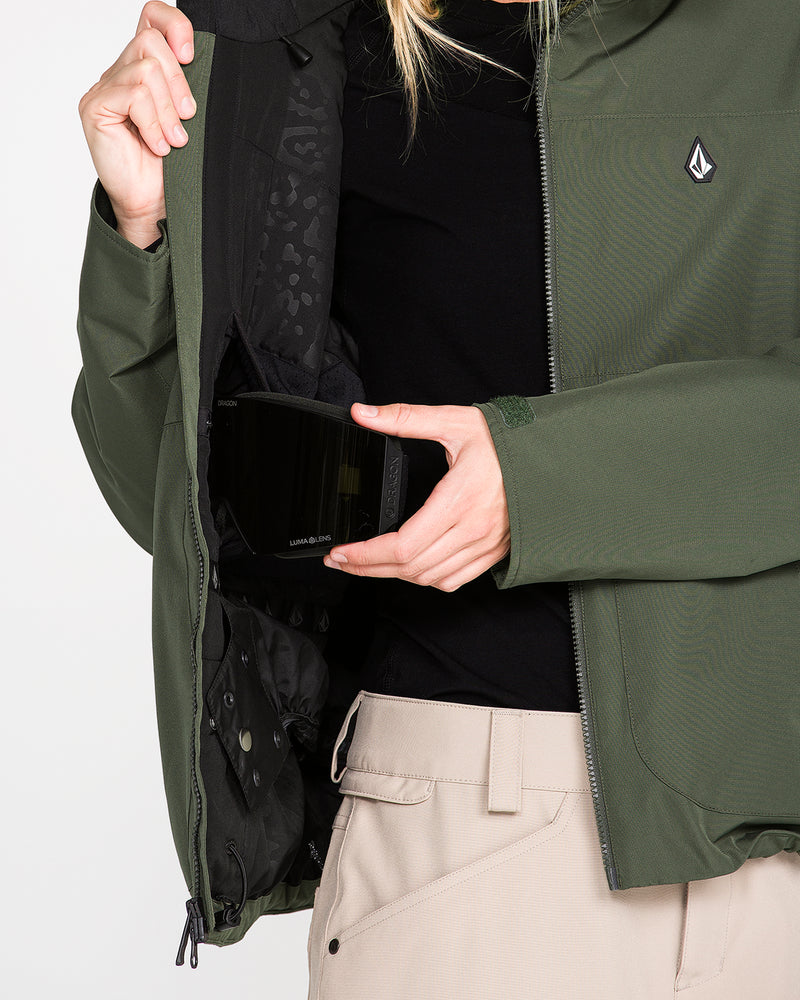 A woman wearing the Volcom Women’s Steele Insulated Jacket in green, featuring recycled insulation, and beige pants displays the inside pocket while holding a pair of ski goggles.