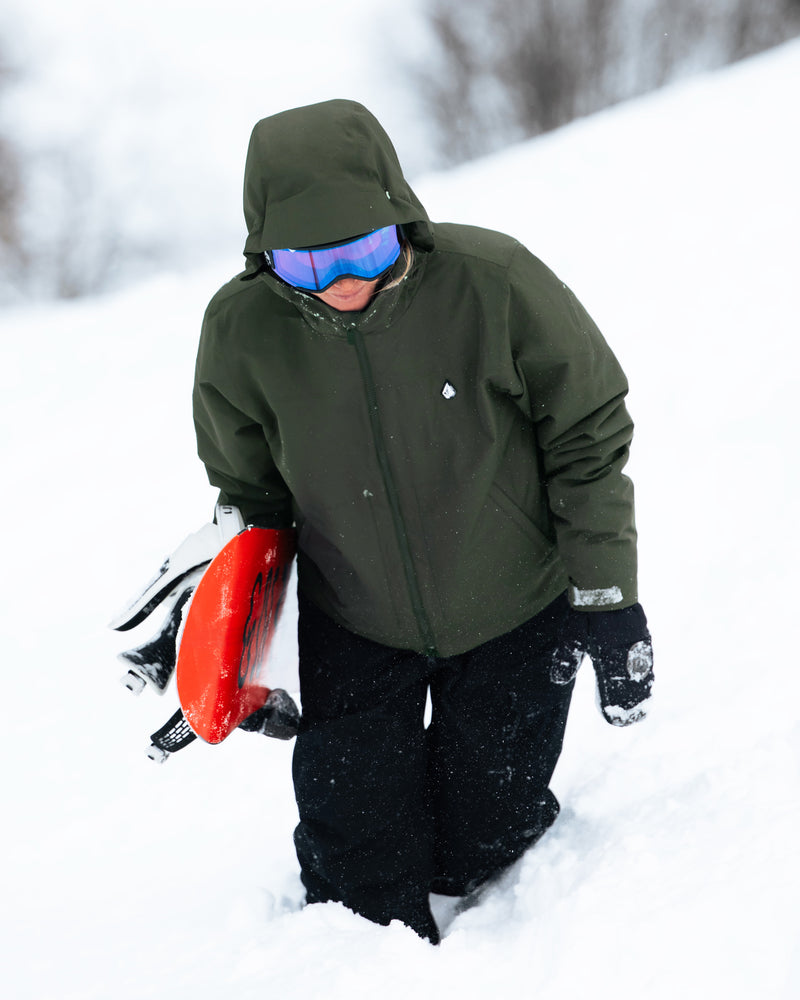 Wearing the Volcom Women’s Steele Insulated Jacket, a person in green with goggles walks uphill in the snow, holding a red sled and carrying a snowboard under one arm.