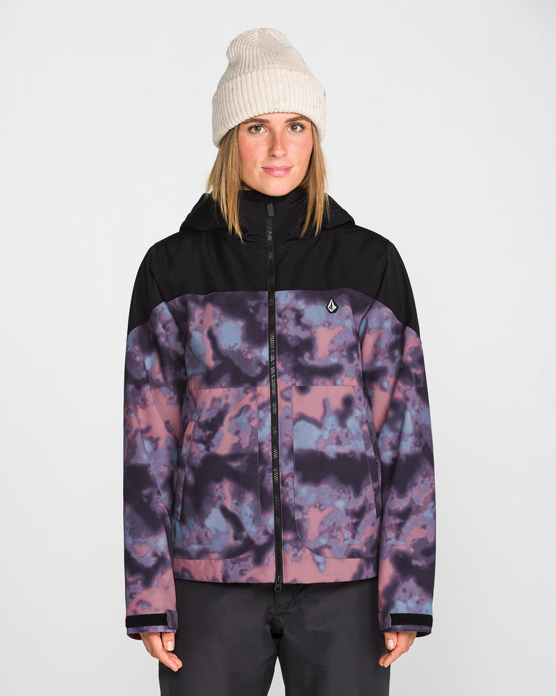 A person wearing a cream knit beanie and the Volcom Women’s Steele Insulated Jacket—featuring a purple-and-black pattern with black shoulders—stands facing forward against a plain light background.