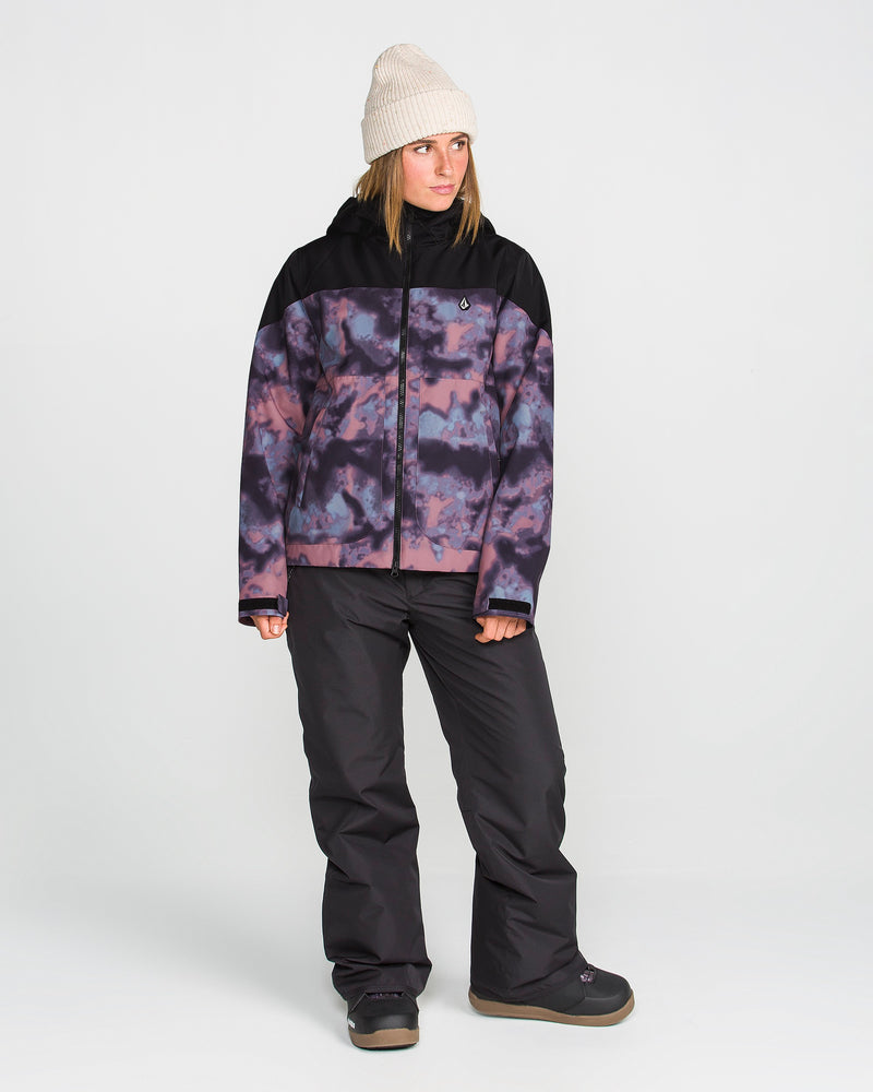 A person wearing a cream beanie, black snow pants, and boots stands against a plain white background, looking to their left. They are dressed in the Volcom Women’s Steele Insulated Jacket—a purple and black patterned women’s jacket made with recycled insulation.