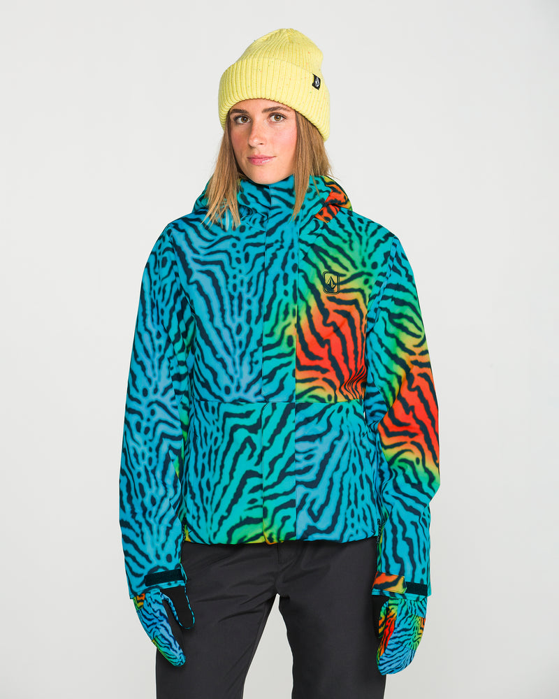 A person wearing the Volcom Women’s Bolt Insulated Jacket in a vibrant, multicolored zebra stripe—engineered with recycled insulation for warmth—paired with matching gloves, black pants, and a yellow knit beanie stands against a plain white background.