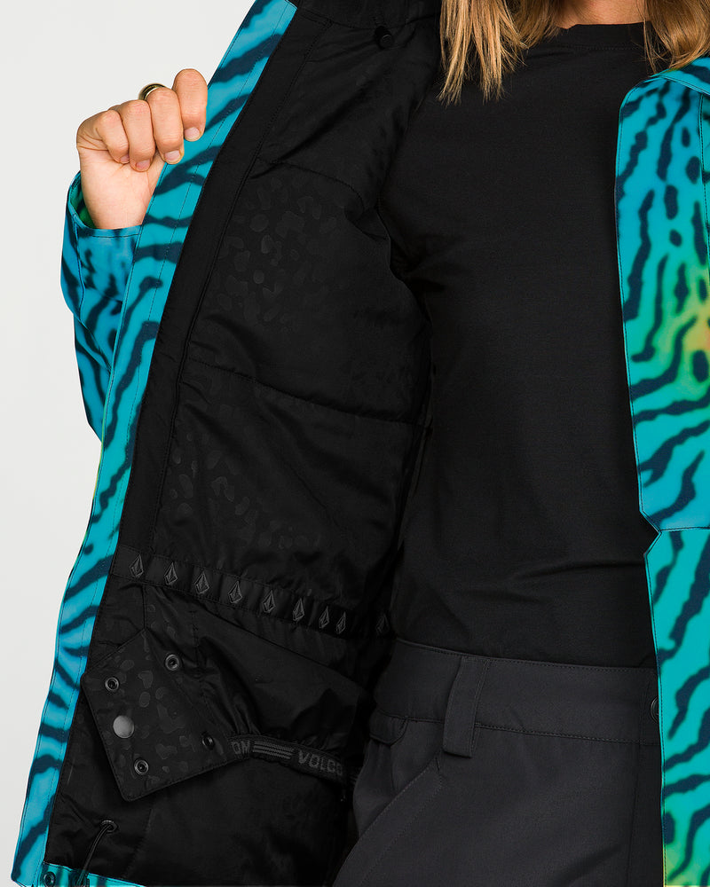 A person wearing the Volcom Women’s Bolt Insulated Jacket in a blue and green animal print style holds it open, revealing the black patterned interior lining. She is dressed in a black shirt and dark pants.