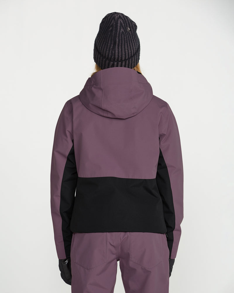 A person is seen from behind against a plain white background, wearing the Volcom Women’s Bolt Insulated Jacket in purple and black, paired with matching pants, a black knit beanie, and gloves.
