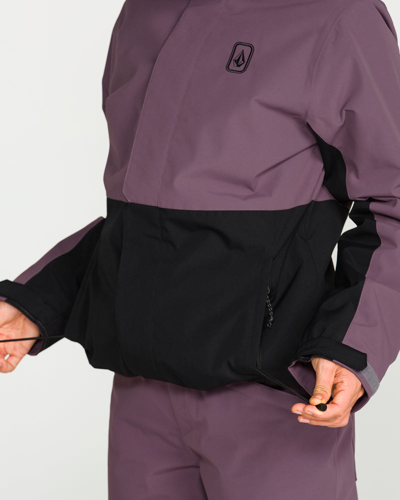 A person wearing the Volcom Women’s Bolt Insulated Jacket in purple and black, along with matching pants, adjusts the waistband while holding a black drawstring.