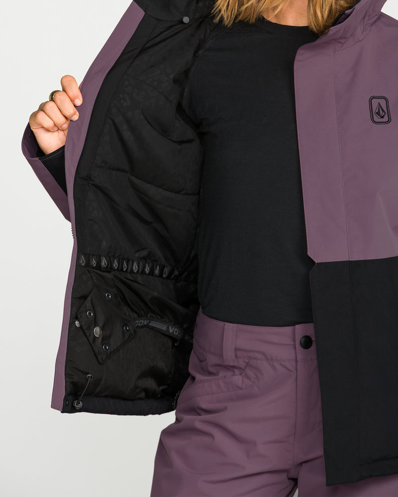 A person wearing a black shirt and purple Volcom Women’s Bolt Insulated Jacket holds open one side of the jacket to reveal its black inner lining. The jacket, featuring a logo on the chest, coordinates with the person’s purple pants.