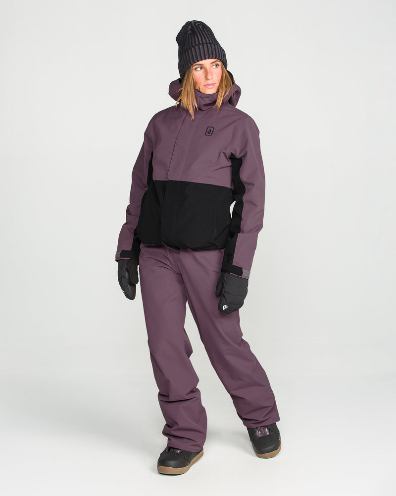 A person wearing the Volcom Women’s Bolt Insulated Jacket in black and purple, along with matching pants, black gloves, dark boots, and a dark beanie stands against a plain white background.