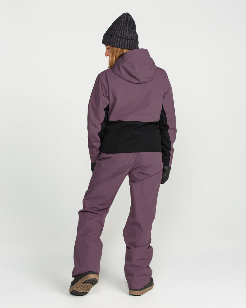 A person stands facing away, wearing the Volcom Women’s Bolt Insulated Jacket in purple and black, paired with matching purple snow pants, a black knit beanie, winter gloves, and brown boots against a plain white background.