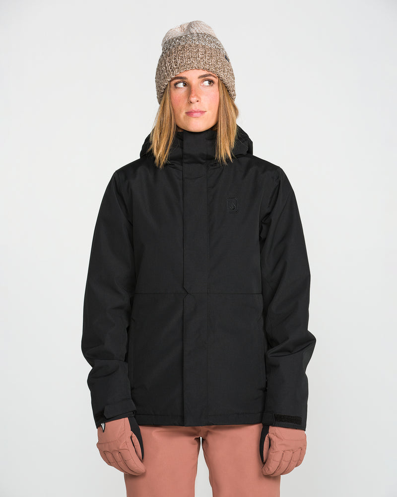 A person wearing the Volcom Women’s Bolt Insulated Jacket in black—ideal for staying warm in cold weather—along with a tan knit beanie and light brown gloves stands facing forward against a plain white background, looking slightly upward and to the side.