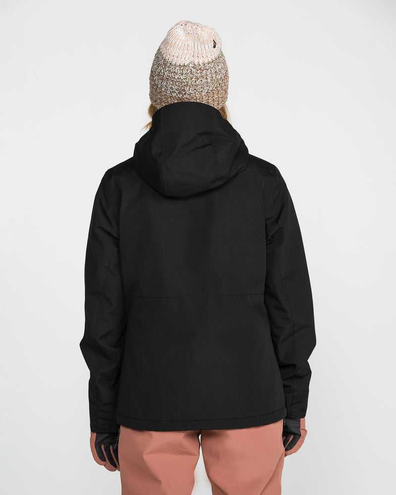 A person stands with their back to the camera against a plain white background, wearing the Volcom Women’s Bolt Insulated Jacket in black, a light knit beanie, and pink pants.