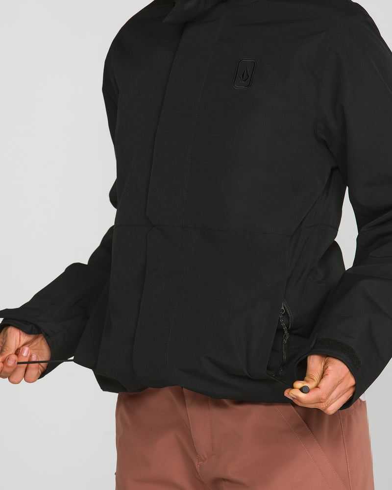 A person wearing the Volcom Women’s Bolt Insulated Jacket in black and brown pants adjusts the jacket’s drawstrings for a customized fit. The image focuses on the torso and hands, with the face not visible.