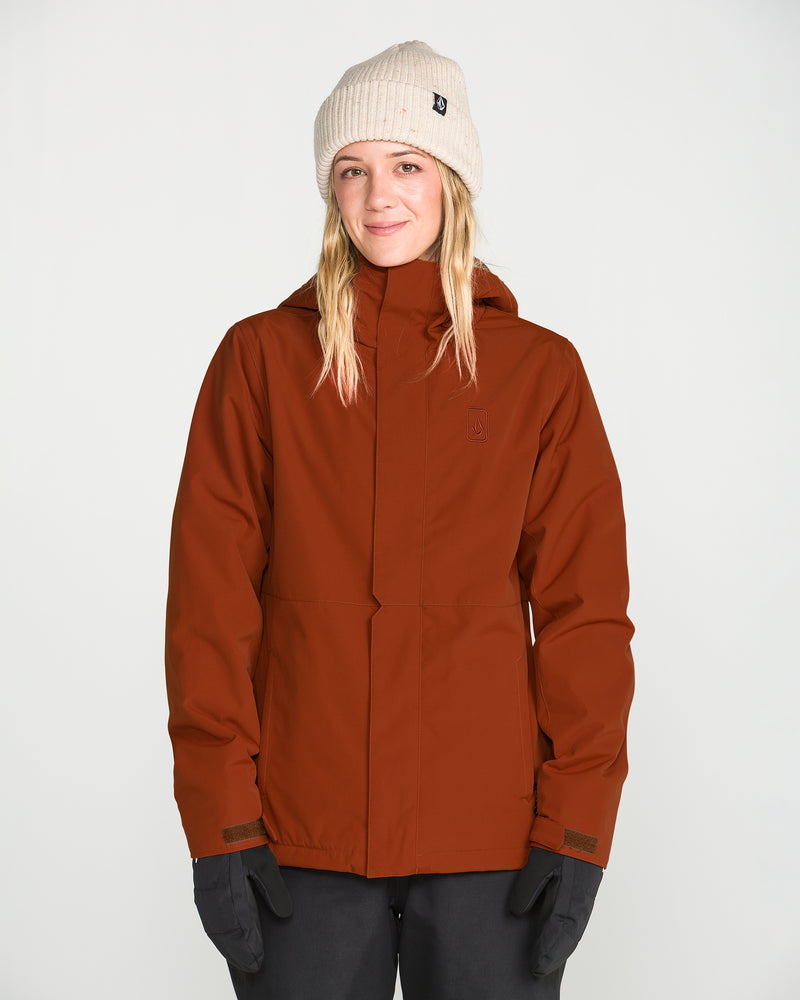 A person wearing a beige knit beanie, black gloves, and dark pants stands against a plain white background, smiling slightly at the camera while modeling the rust-colored Women’s Bolt Insulated Jacket by Volcom.