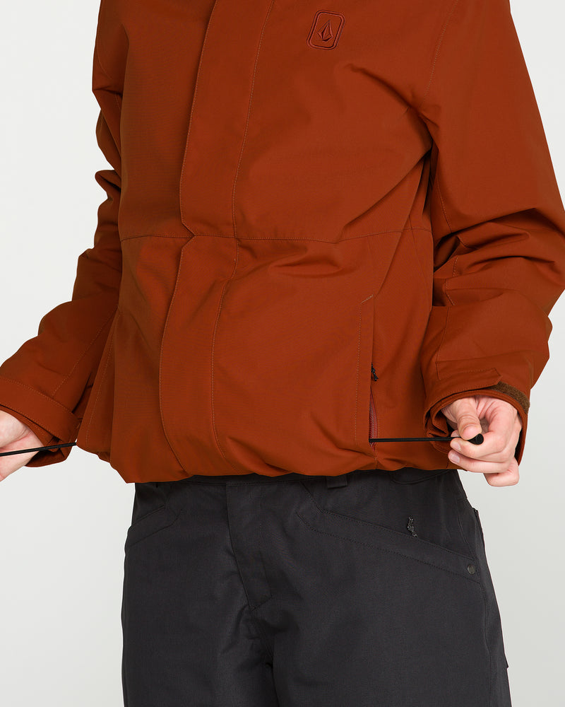 A person wearing the Volcom Women’s Bolt Insulated Jacket in a rust hue and black pants pulls on the jacket’s adjustable waist drawstring. This stylish piece features recycled insulation and a PFAS-free DWR finish. The person’s head is not visible in the image.