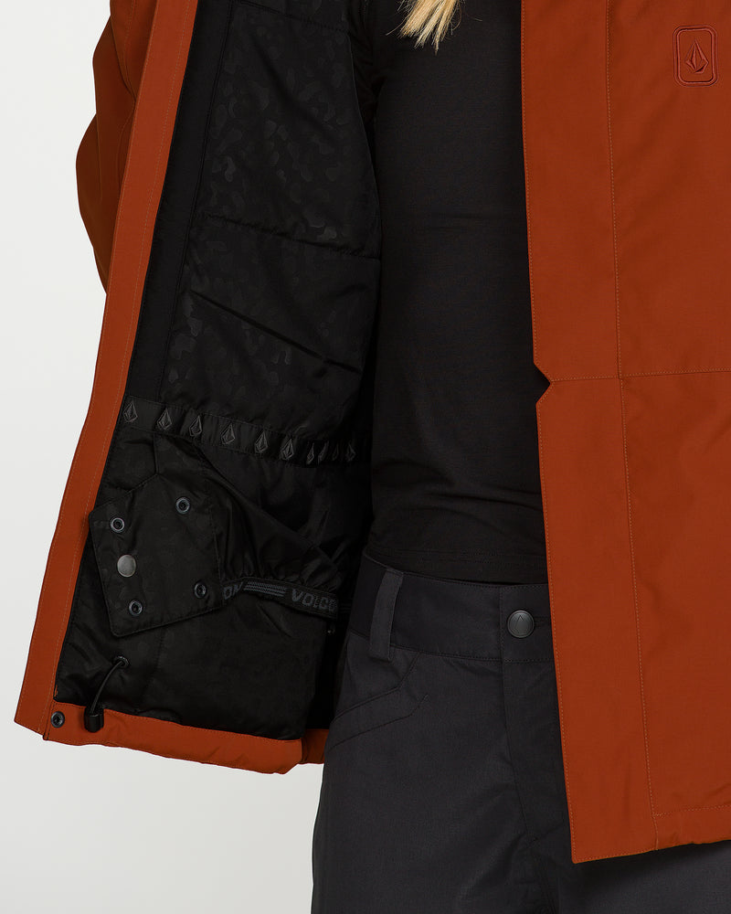 A person with blond hair, wearing Volcom’s Women’s Bolt Insulated Jacket in burnt orange and dark pants, opens the jacket to reveal its black, patterned interior with an inside pocket—highlighting its recycled warmth. Part of a black shirt is also visible.