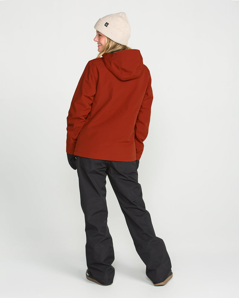 Wearing the Volcom Women’s Bolt Insulated Jacket in red, paired with black snow pants and gloves, a beige beanie, and brown shoes, a person stands against a plain white background. They face away while looking over their shoulder and smiling.
