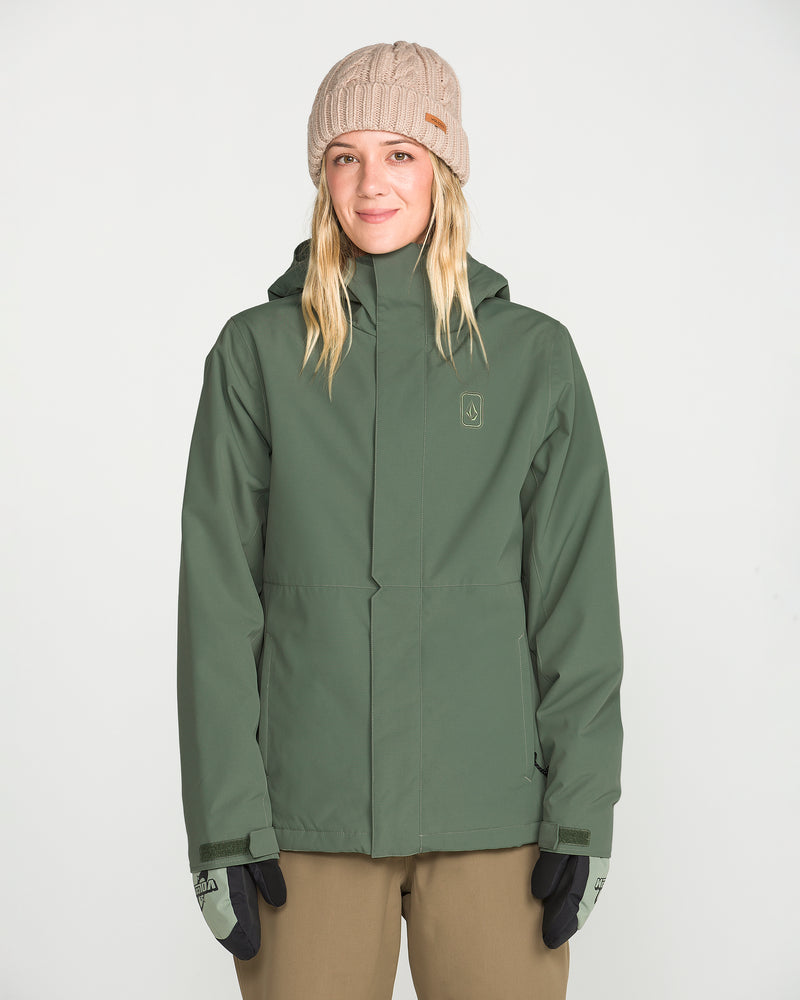 A woman stands against a plain white background, facing the camera and smiling slightly, dressed in a Volcom Women’s Bolt Insulated Jacket in green with V-Science 2-Layer technology, a light brown knit beanie, black gloves, and tan pants.