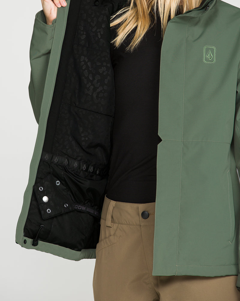 A person wearing the Volcom Women’s Bolt Insulated Jacket in green and brown pants opens the jacket to reveal its black, leopard-print lining, crafted with V-Science 2-Layer technology for recycled warmth. Only the lower half of their face and torso are visible, highlighting the sustainable style of this Volcom outerwear.