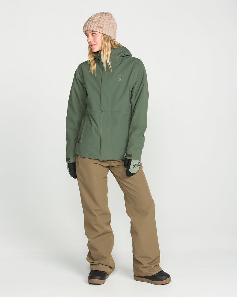 Wearing a green Volcom Women’s Bolt Insulated Jacket—featuring V-Science 2-layer recycled warmth—paired with tan pants, black gloves, black boots, and a beige beanie, a person stands against a plain white background, glancing to the side with a slight smile.