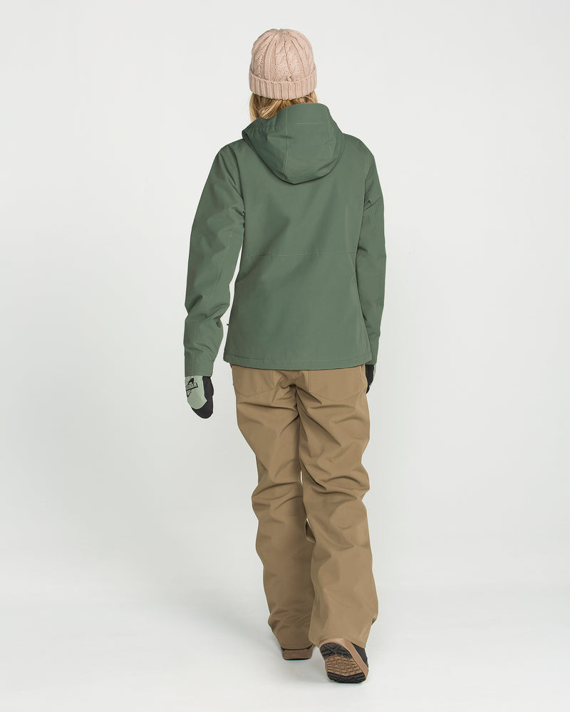 A person stands with their back to the camera against a plain white background, wearing a green Volcom Women’s Bolt Insulated Jacket made with V-Science 2-Layer technology for recycled warmth, paired with tan snow pants, gloves, and a beige knit hat.