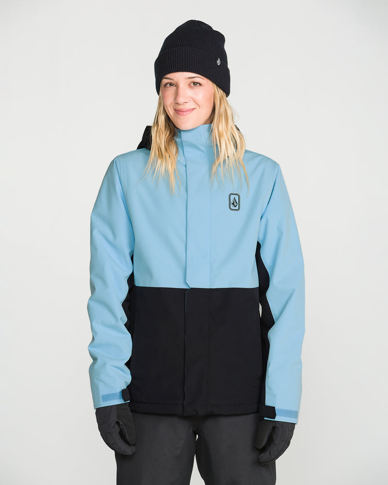 A person with long hair wearing a black beanie, black gloves, and dark pants stands in front of a plain light background, smiling slightly while modeling the Volcom Women’s Bolt Insulated Jacket—a light blue and black women’s jacket designed for recycled warmth.