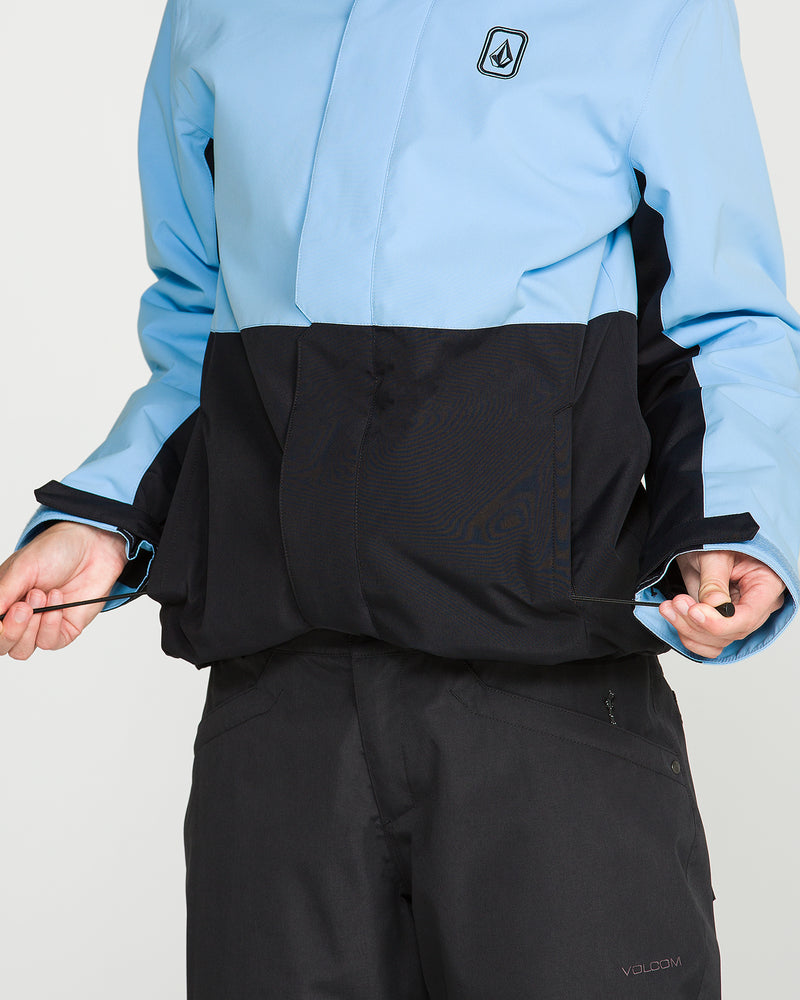A person wearing the Volcom Women’s Bolt Insulated Jacket in light blue and black, along with black pants, pulls on the jacket’s waist drawstrings; only their torso and hands are visible.