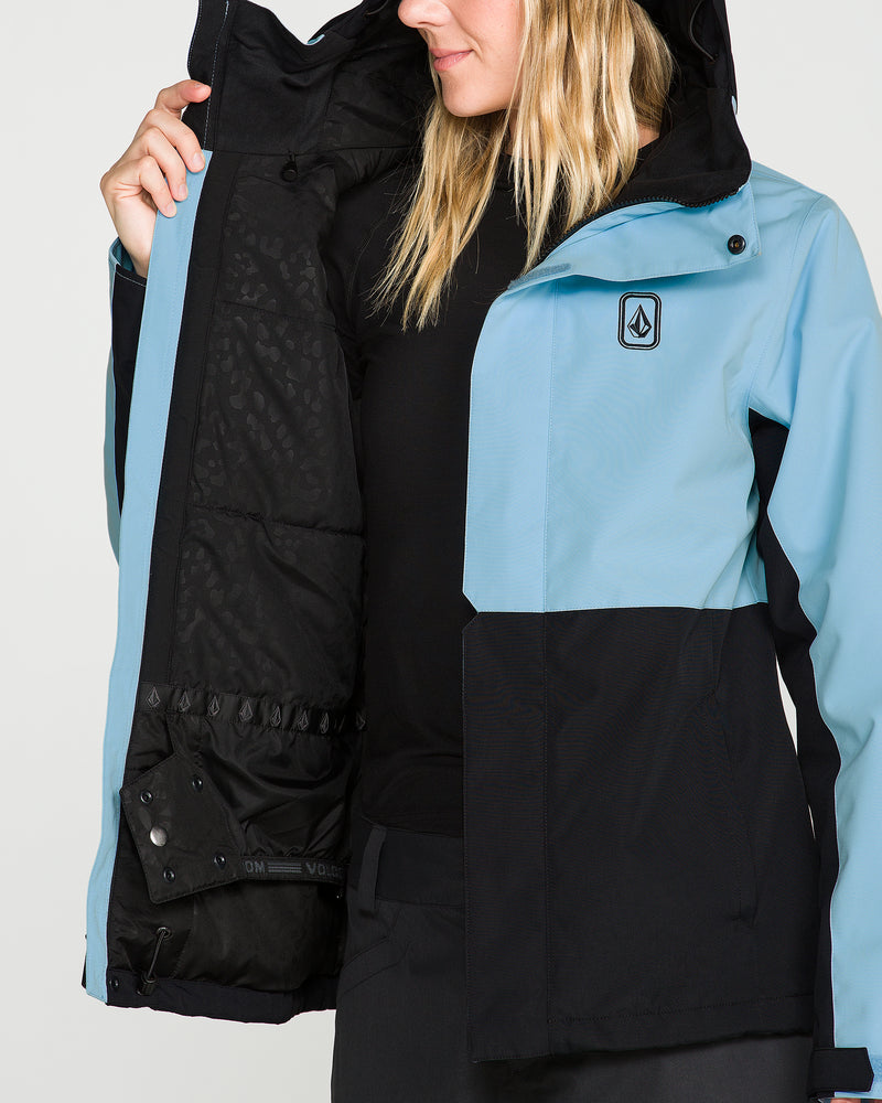 A woman holds open the Volcom Women’s Bolt Insulated Jacket in blue and black, displaying its inner lining, pockets, and snaps. With her face partially hidden by loose blonde hair, she emphasizes the jacket’s practical features—showcasing a design made for recycled warmth.