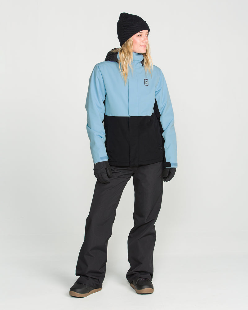 A person is shown standing sideways against a plain white background, wearing the Volcom Women’s Bolt Insulated Jacket in blue and black, paired with black pants, gloves, a beanie, and boots.