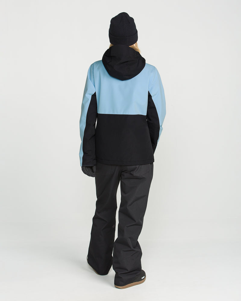A person is standing facing away, wearing a black beanie and the Volcom Women’s Bolt Insulated Jacket—blue and black, designed for recycled warmth—paired with black snow pants and gloves, set against a plain white background.