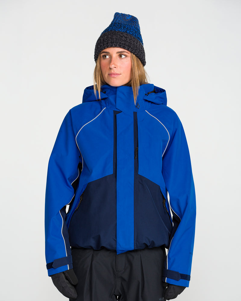 A person is wearing the Volcom Women’s Unisex Yourfav Baggy Gore Jacket in blue, paired with dark pants, black gloves, and a blue knit beanie. They are standing against a plain light background and looking slightly to the side.