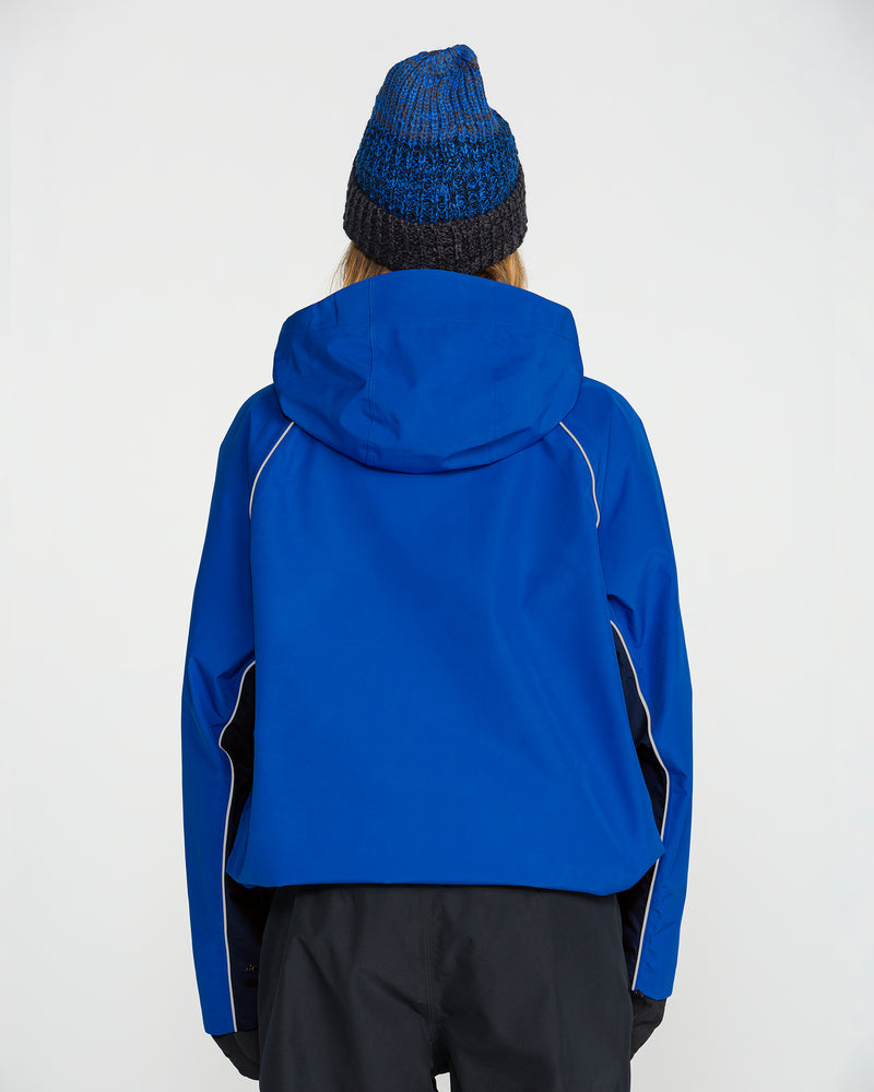 A person is shown from behind against a plain white background, wearing the Volcom Women’s Unisex Yourfav Baggy Gore Jacket in blue with a hood, paired with a blue knit hat.