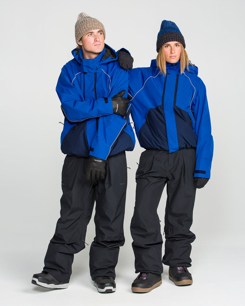 Two people are wearing Volcom Women’s Unisex Yourfav Baggy Gore Jackets in blue, paired with black snow pants, gloves, and knitted hats. Standing together against a plain white background, one rests their arm on the other’s shoulder as they both face forward.