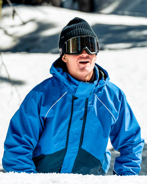 A person wearing the Volcom Women’s Yourfav Baggy Gore Jacket in blue, along with a black hat and ski goggles, stands outside on a snowy day, looking upward.