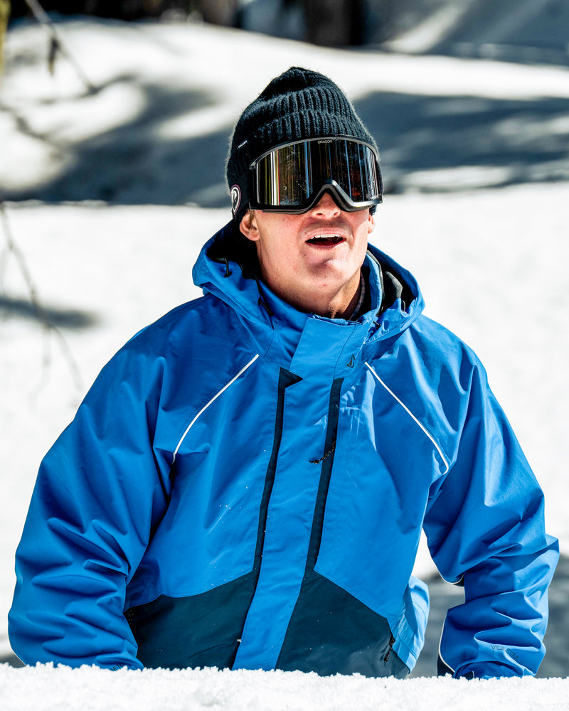 A person wearing the Volcom Women’s Yourfav Baggy Gore Jacket in blue, along with a black hat and ski goggles, stands outside on a snowy day, looking upward.
