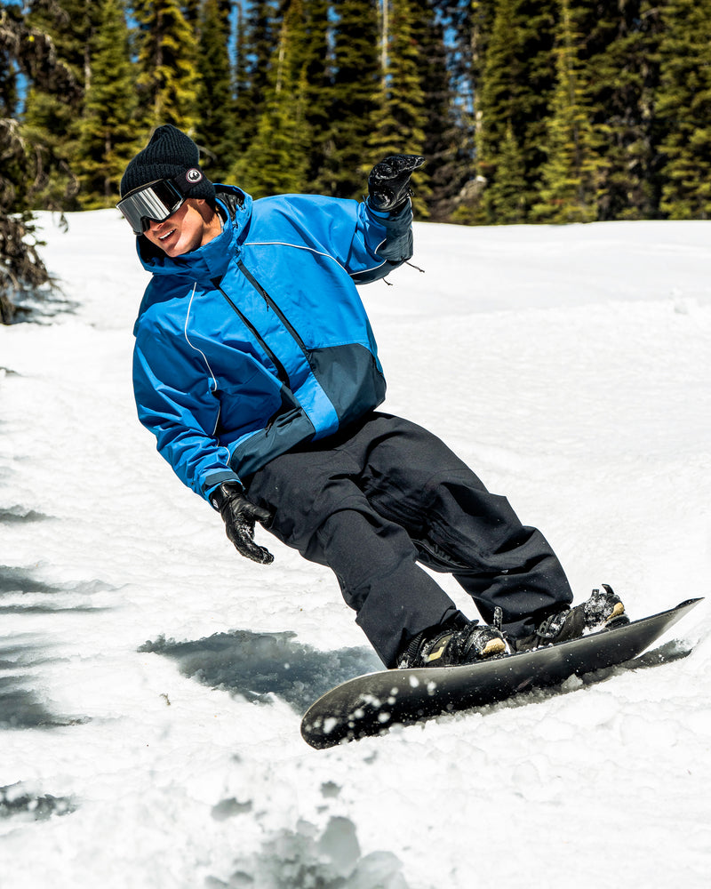 Wearing the Volcom Women’s Yourfav Baggy Gore Jacket in blue and black pants, a snowboarder carves down a snowy slope bordered by evergreen trees beneath a sunny sky.